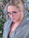 Four-eyed blonde Alexis Malone in smart office suit gives a close-up of her pink slit